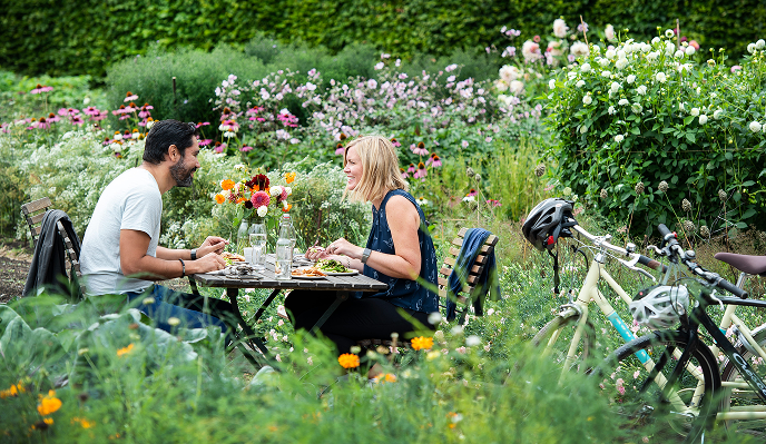 En man och en kvinna äter en måltid tillsammans vid ett bord i en grönskande trädgård, med två cyklar parkerade intill.