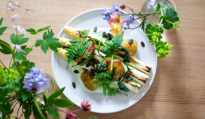 En vackert dukad tallrik med sommarsäsongens råvaror som vit sparris och potatis intill snittblommor i vasar.